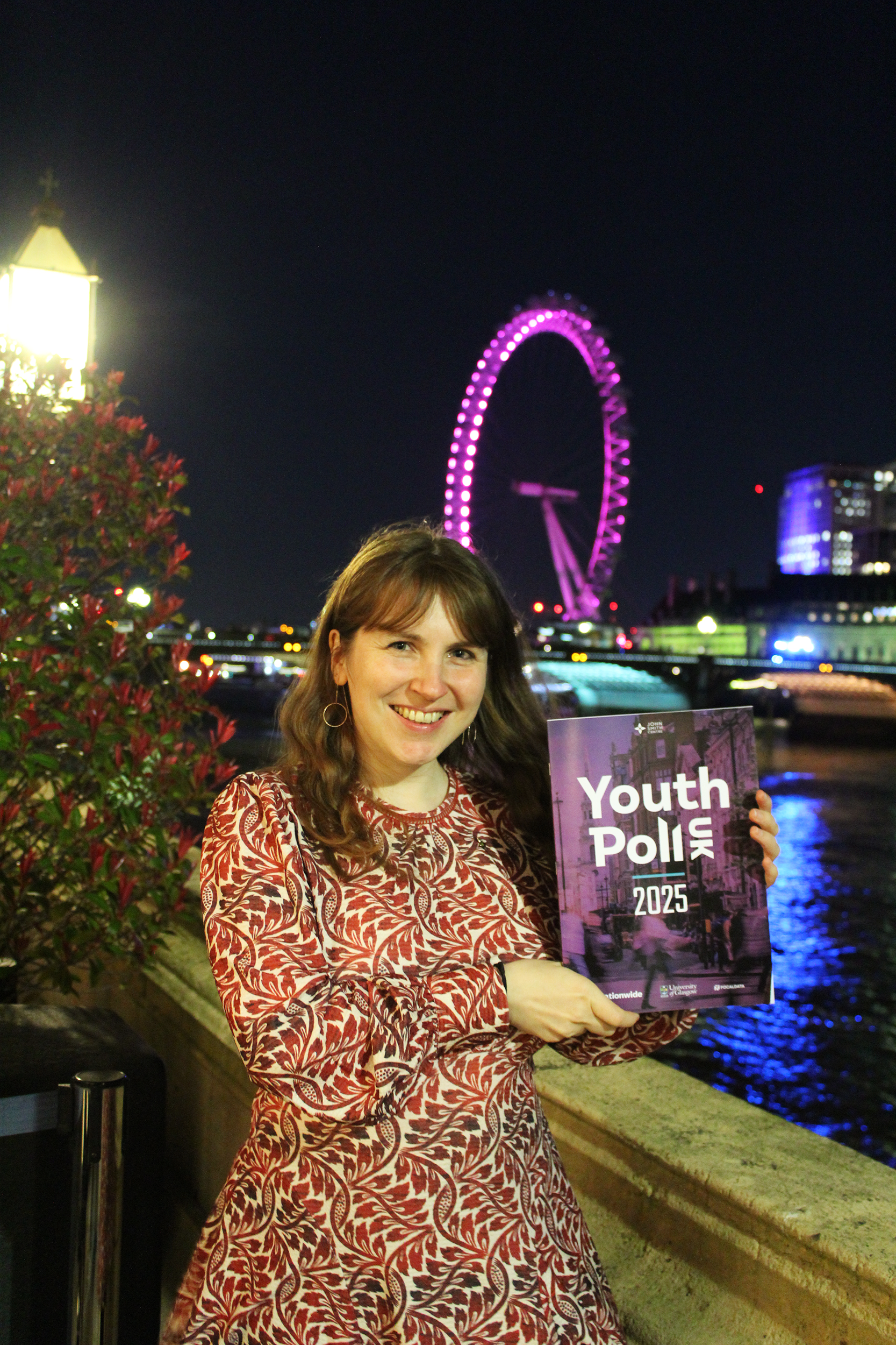 A photo of Dr Elisabeth Loose standing outside at night, holding up a copy of the UK Youth Poll 2025 report. The River Thames and the London Eye are in the background. Elli has long, wavy brown hair, blue eyes, and is smiling.