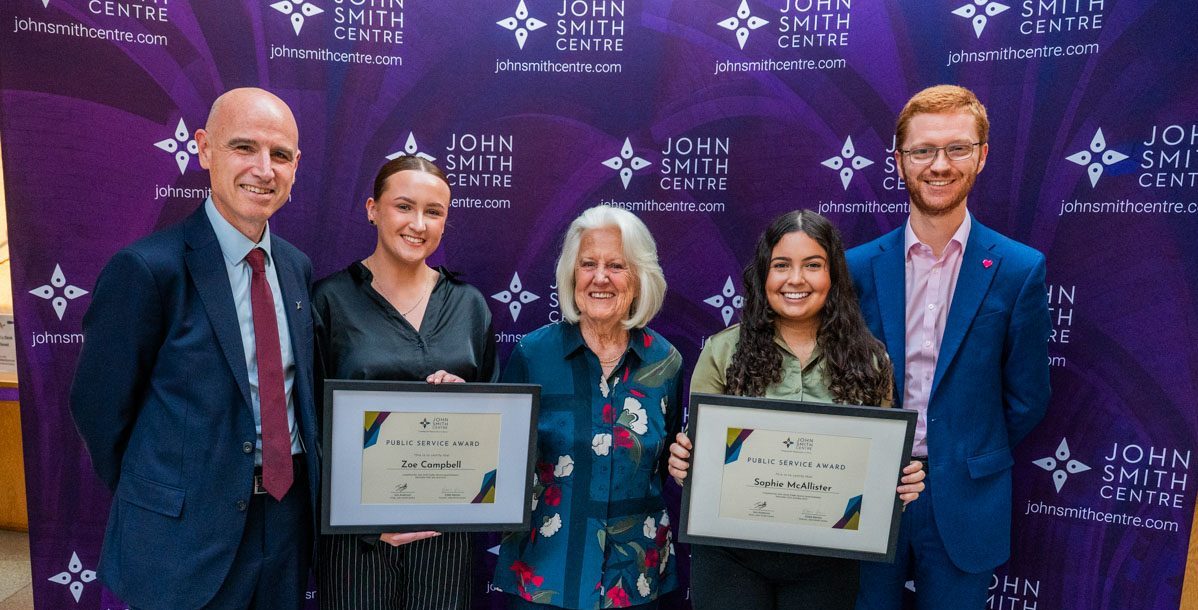 John Smith Public Service Award recipients Zoe Campbell and Sophie McAllister with John Smith Centre Director Eddie Barnes, Baroness Elizabeth Smith, and Ross Greer MSP at the 2025 John Smith Centre Annual Reception at the Scottish Parliament Building, Edinburgh, September 2025. Zoe and Sophie are holding framed certificates. Behind the group is a large John Smith Centre banner.
