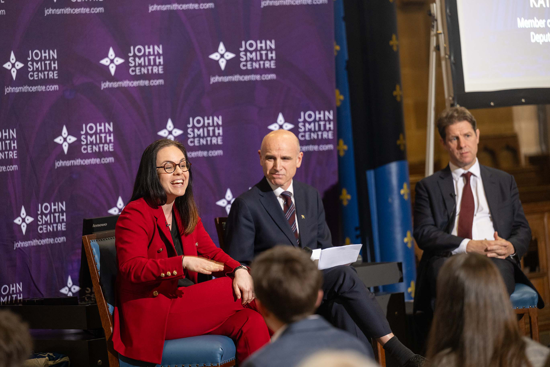 Kate Forbes MSP, Eddie Barnes and Fraser Nelson sat in chairs onstage in front of a seated crowd in the University of Glasgow's Bute Hall for the 2026 Surviving Politics event, February 2026. There is a banner behind them displaying the John Smith Centre logo.