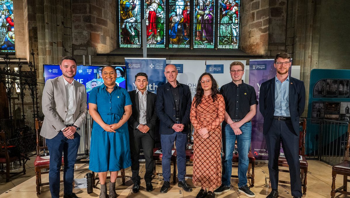 Participants standing onstage at the John Smith Centre's Scottish Hustings for Young People in St Giles' Cathedral, Edinburgh. L-R: Ross Hutton (Scottish Conservatives), Iris Duane (Scottish Green Party), Jamie McGuire (Reform UK), Eddie Barnes (John Smith Centre Director), Eleanor Ryan-Saha (Scottish Labour), Callum Cox (SNP), Daniel Khan-O'Malley (Scottish Liberal Democrats).
